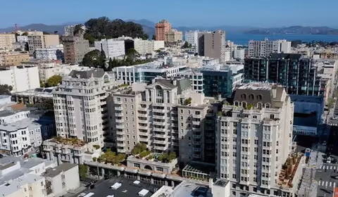 Aerial view of a large residential building complex in an urban area with multiple mid-rise buildings, greenery on terraces, and a distant view of water and hills under a clear sky.