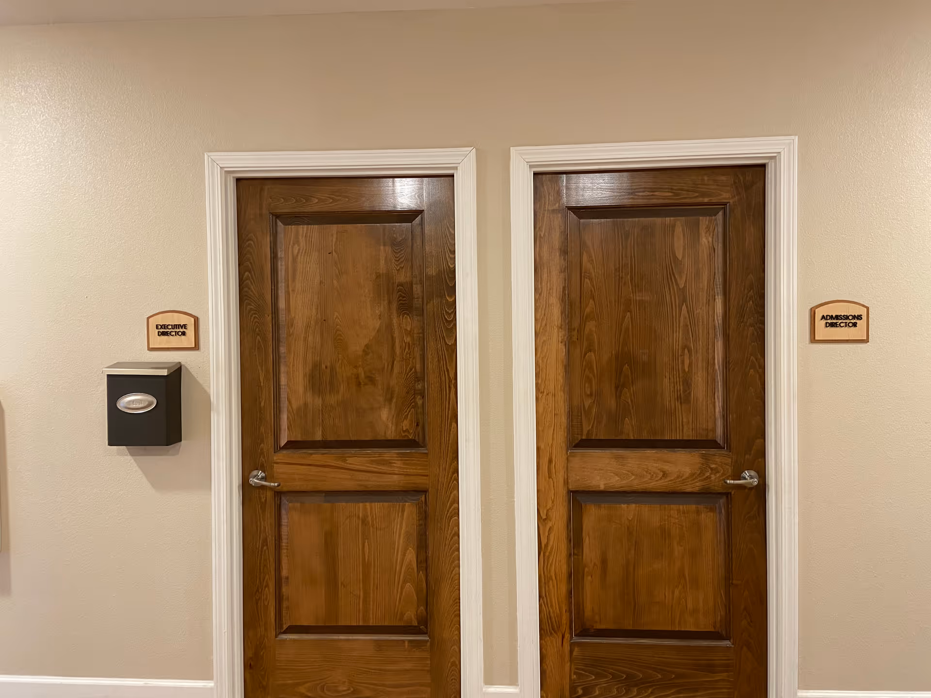 Two closed wooden doors with white trim in a beige hallway. The door on the left has a sign above it that reads 'EXECUTIVE DIRECTOR' and a black box mounted on the wall beside it. The door on the right has a sign above it that reads 'ADMISSIONS DIRECTOR'.