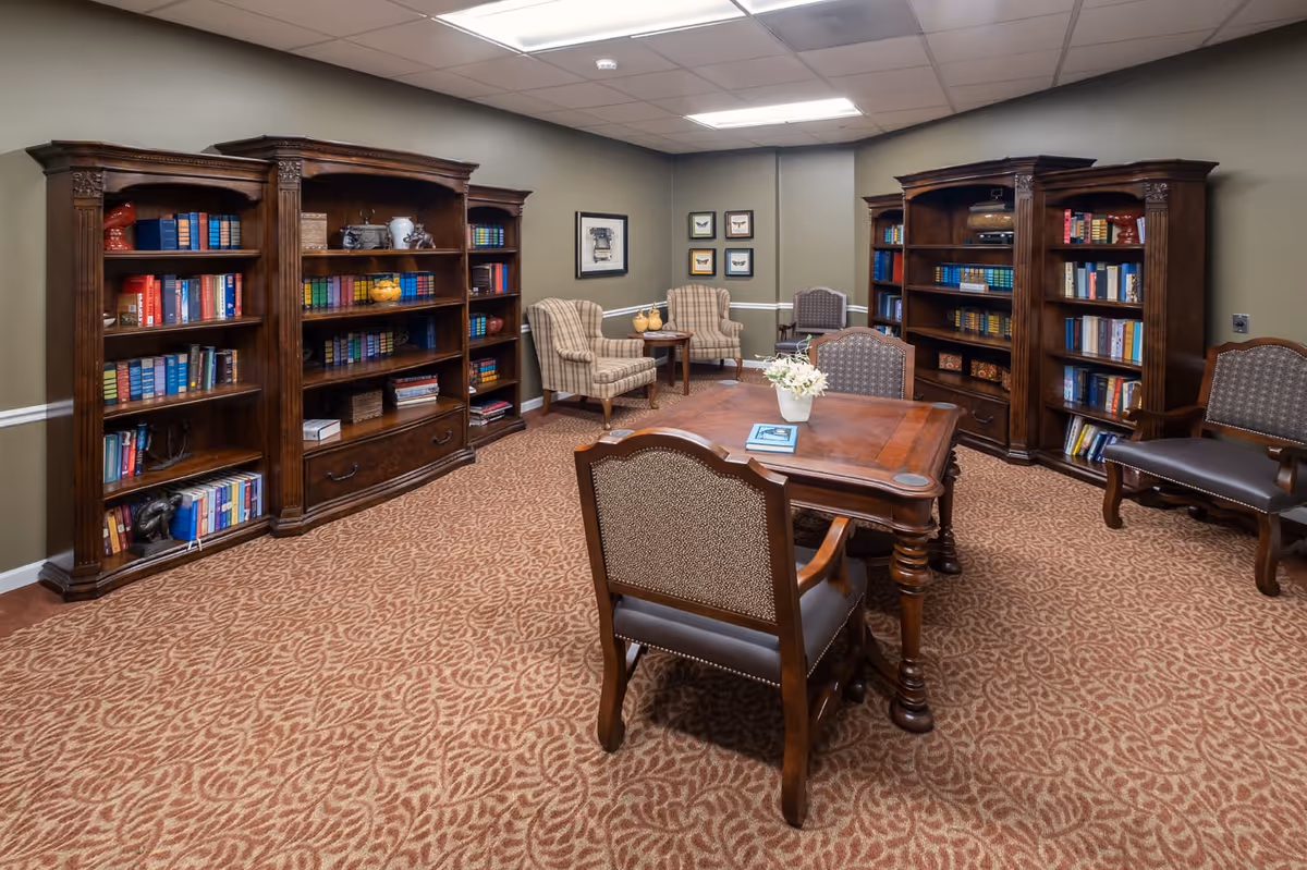 A cozy library room with three large wooden bookshelves filled with books and decorative items. There is a wooden table with a flower vase and a book on it, surrounded by four upholstered chairs. In the corner, there are two plaid armchairs with a small round table between them. The walls are decorated with framed pictures, and the carpet has a leaf pattern.