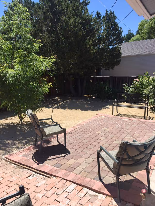 Sunny backyard patio with brick pavers, metal chairs and a bench surrounded by trees and shrubs.