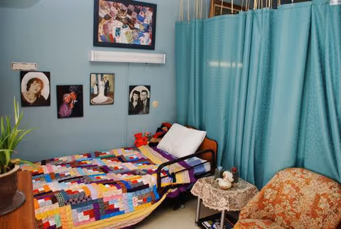 A small bedroom with a single bed covered by a colorful patchwork quilt. The bed has a white pillow and a black bed rail. Next to the bed is a small round table with a lace tablecloth, a teapot, and a cup. An upholstered chair with an orange and beige floral pattern is beside the table. The walls are light blue and decorated with several framed photographs and artwork. A teal privacy curtain hangs near the bed, partially enclosing the space.