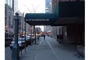 Sidewalk view of the Mary Manning Walsh Nursing Home entrance with a green awning displaying the facility name. The street is lined with parked cars and buildings, with a few pedestrians walking along the sidewalk.