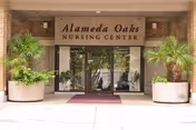 Entrance to Alameda Oaks Nursing Center with glass double doors, a maroon welcome mat, and large potted plants on either side under a brick facade.