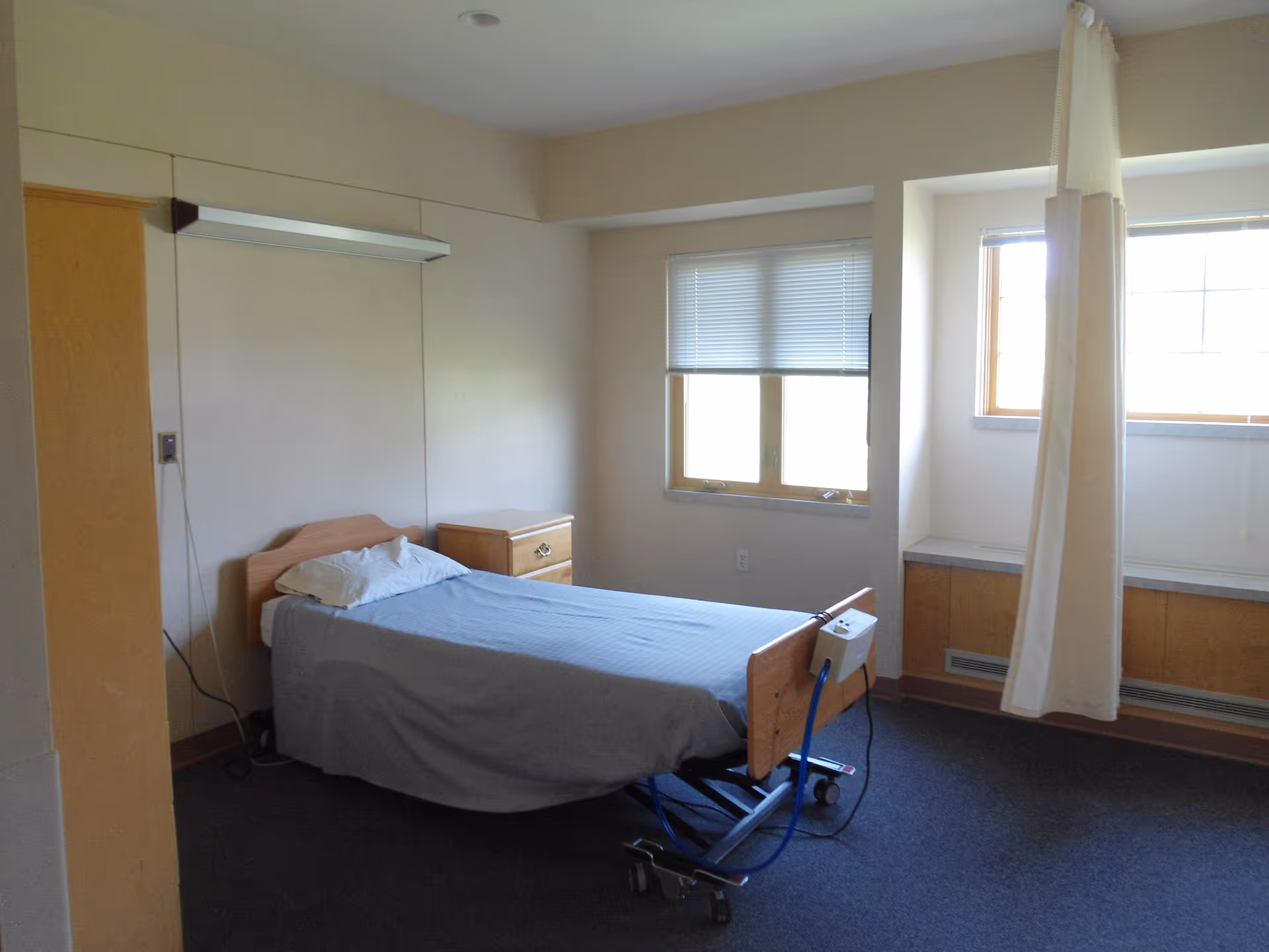 A sparse patient bedroom with an adjustable hospital bed on wheels, a nightstand, windows with blinds, and a privacy curtain.