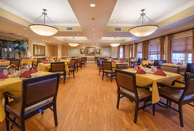 Spacious dining room with multiple tables covered in yellow tablecloths and set with red folded napkins, glasses, and silverware. The room has wooden flooring, large windows with brown curtains, and ceiling lights providing warm illumination. There are paintings on the far wall and a large plant in the corner.