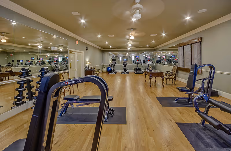 A fitness room with wooden floors and beige walls, equipped with various exercise machines including weight benches, dumbbells, and cardio equipment. Large mirrors cover one wall, reflecting the room and equipment. Ceiling fans and recessed lighting illuminate the space.