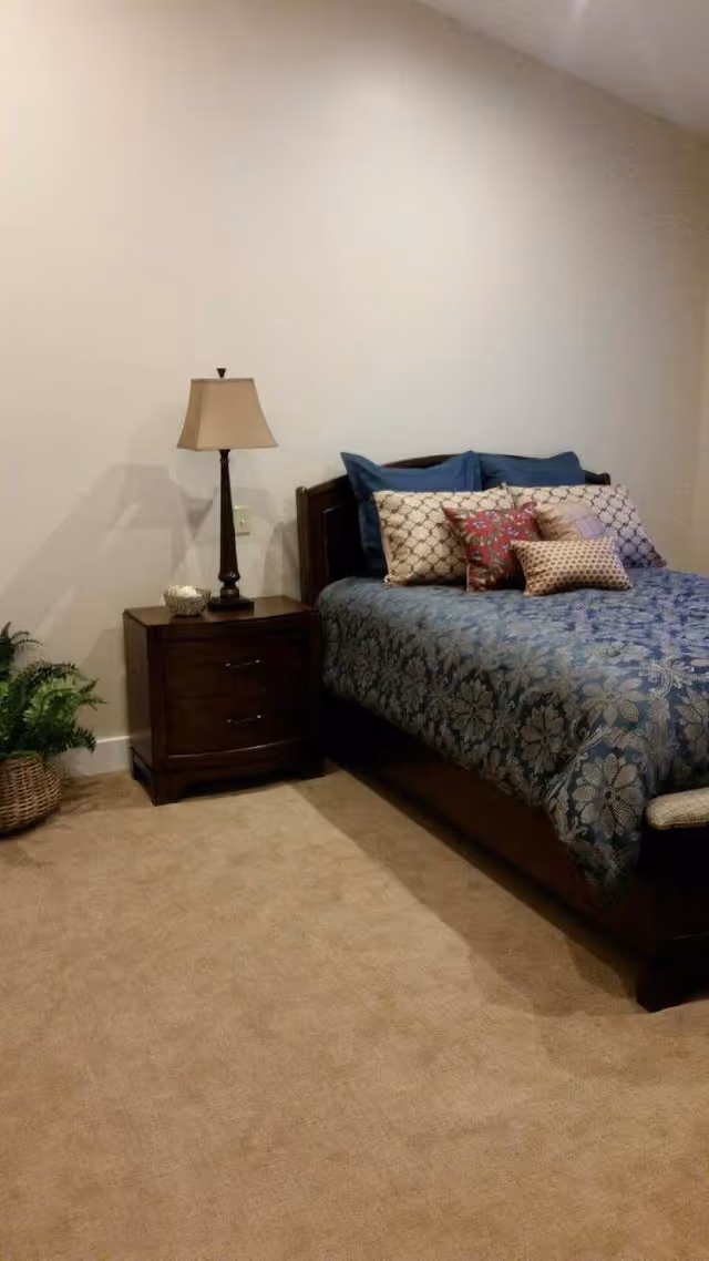 A bedroom with a wooden bed frame and a blue floral bedspread. The bed is adorned with multiple decorative pillows in various patterns and colors. Next to the bed is a wooden nightstand with two drawers, a table lamp with a beige shade, and a small decorative item. There is a woven basket with a green plant on the floor beside the nightstand. The walls are plain and light-colored, and the floor is carpeted in a neutral tone.
