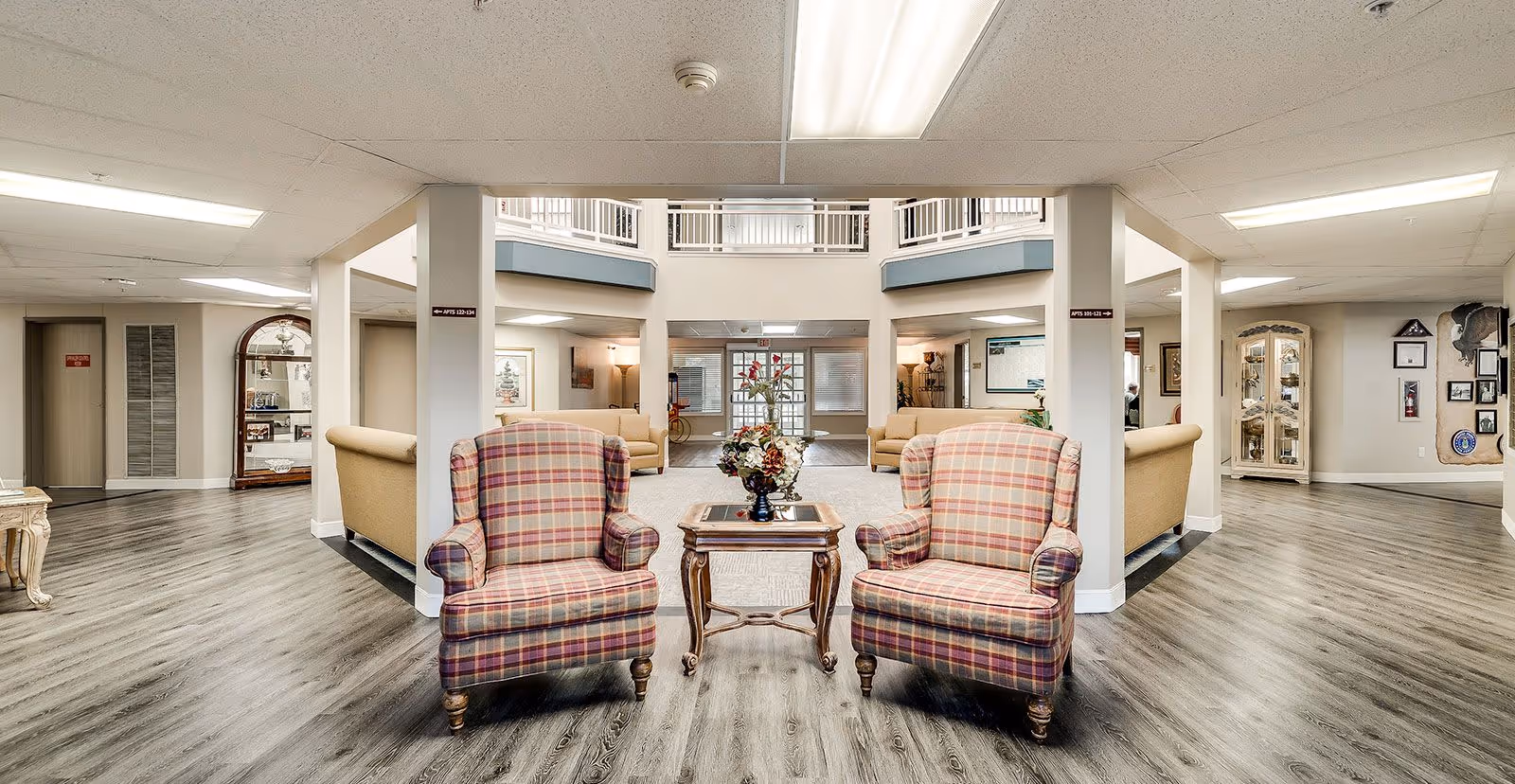 A spacious and well-lit common area in a senior living facility featuring two plaid upholstered armchairs facing a small wooden table with a floral arrangement. The room has wood flooring, beige walls, and multiple seating areas with sofas in the background. The ceiling has fluorescent lighting, and there are decorative elements such as framed pictures and a glass display cabinet along the walls.