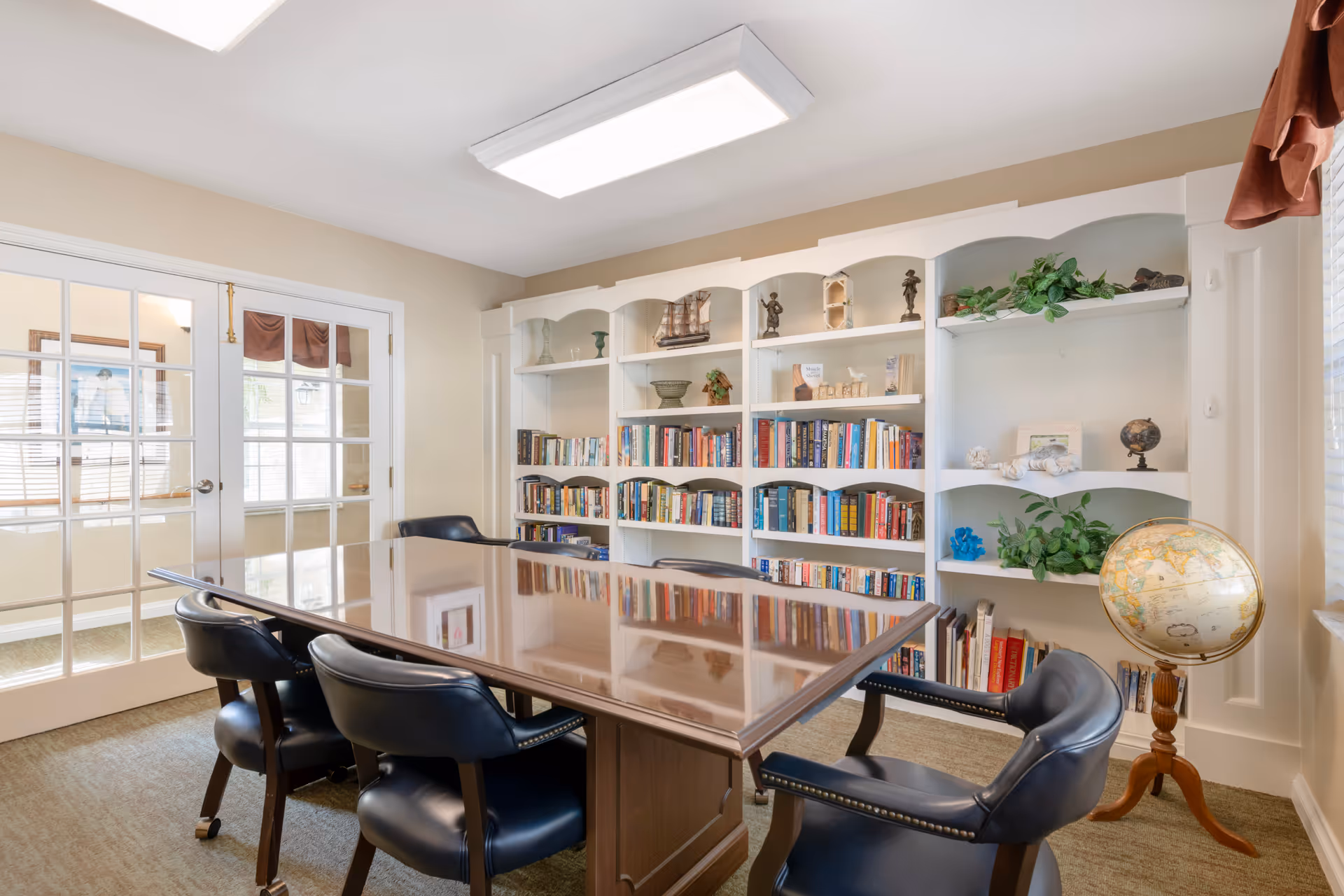 A well-lit room with a large wooden table surrounded by six black leather chairs on wheels. Behind the table is a white built-in bookshelf filled with books and decorative items, including plants, a globe, and small sculptures. The room has beige walls, a carpeted floor, and a set of French doors with glass panes. A globe on a wooden stand is positioned near the window with blinds partially open.