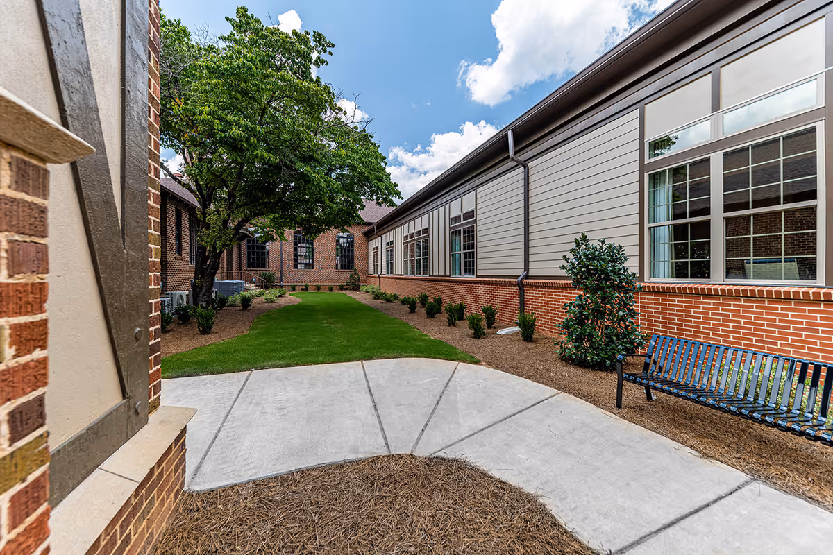 Outdoor courtyard area of Alexander IV Senior Living with a concrete pathway, green grass, small bushes, a large tree, and a black metal bench next to a building with brick and siding exterior under a partly cloudy blue sky.