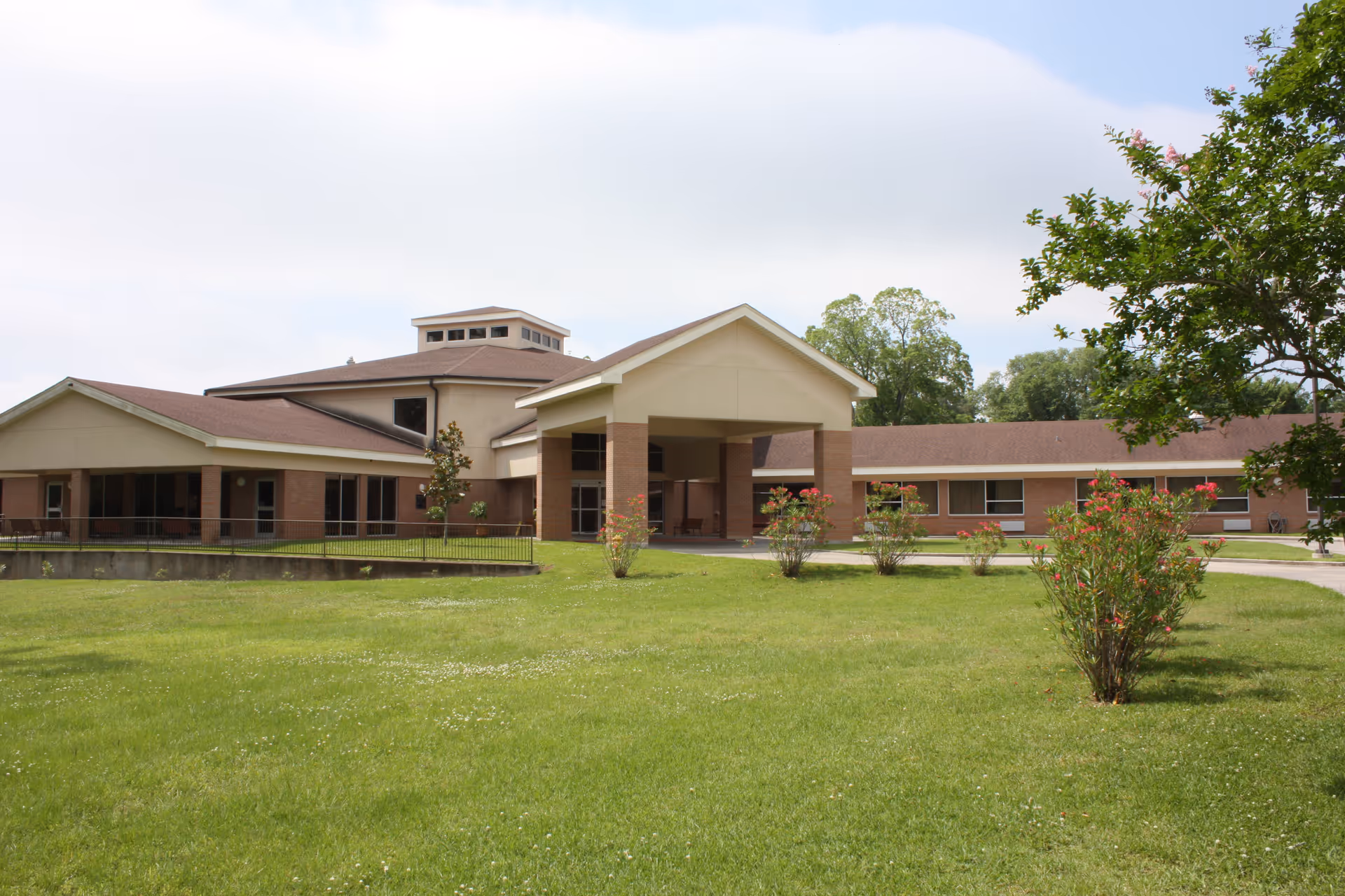 Front exterior view of a single-story nursing facility and independent living center building with a covered entrance, surrounded by a well-maintained lawn and small flowering bushes under a partly cloudy sky.