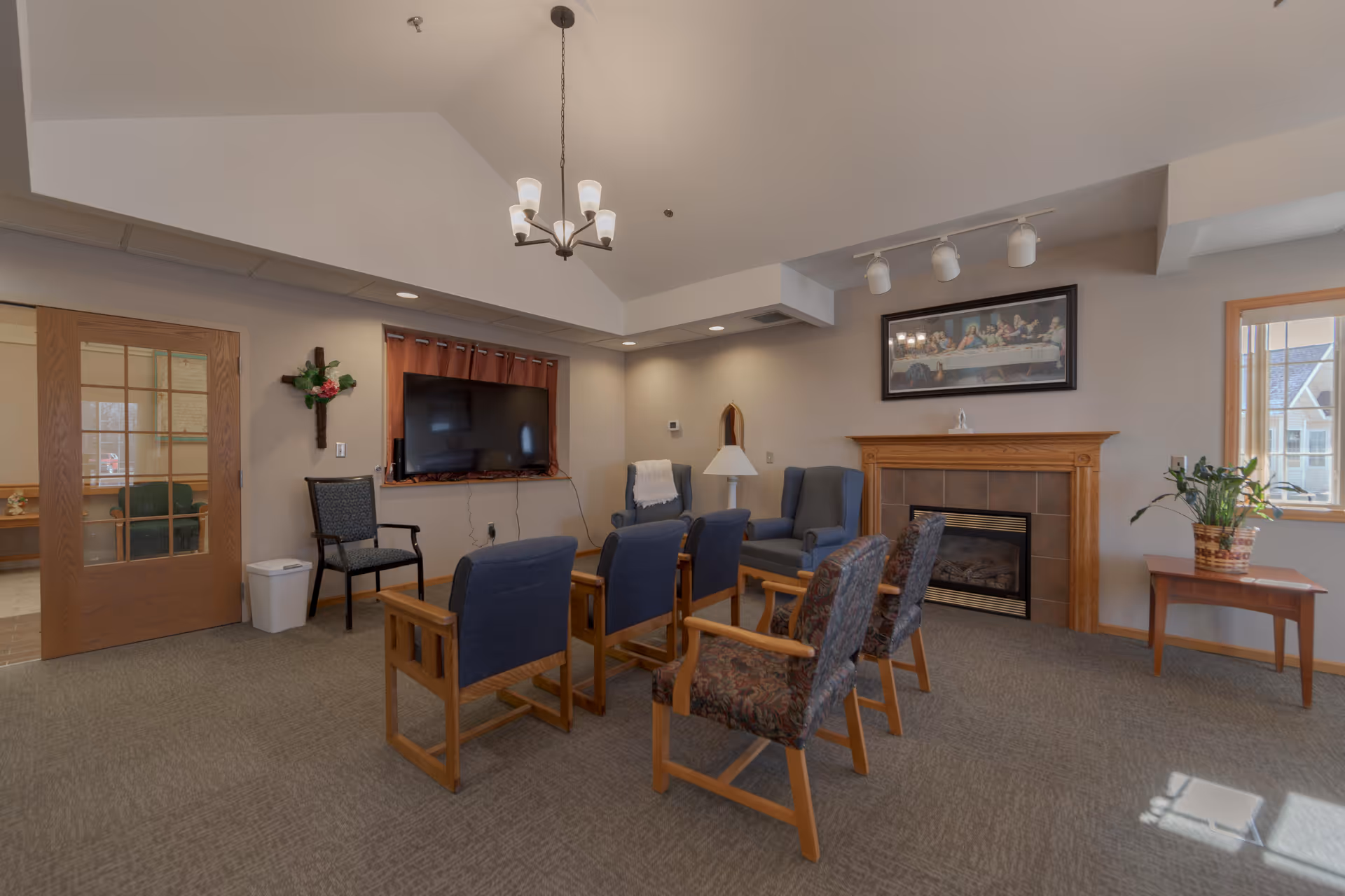 A cozy common area with multiple chairs arranged around a small wooden table, a wall-mounted flat-screen TV, a fireplace with a wooden mantle, a framed picture above the fireplace, a potted plant on a side table near a window, and a chandelier hanging from the ceiling.
