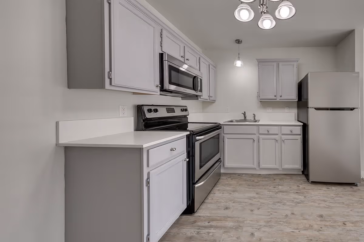 A modern kitchen with light gray cabinets, a stainless steel refrigerator, stove, and microwave. The kitchen has white countertops, a double sink, and wood-patterned flooring. There is a ceiling light fixture with multiple bulbs and a hanging pendant light above the sink area.