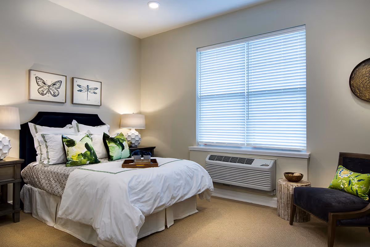A neatly made bed with white and patterned bedding and green accent pillows in a bedroom. Two bedside tables with lamps flank the bed. Above the bed are two framed pictures of a butterfly and a dragonfly. A window with closed blinds is on the right wall above an air conditioning unit. A dark upholstered chair with a green pillow and a small wooden side table with a decorative bowl are also visible.