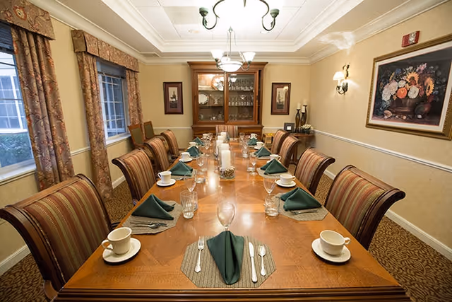 A formal dining room with a long wooden table set for twelve people. Each place setting includes a white cup and saucer, a glass, silverware, and a green folded napkin on a placemat. The room has patterned curtains on the windows, a wooden china cabinet with glassware, framed artwork on the walls, and warm lighting from ceiling fixtures and wall sconces.