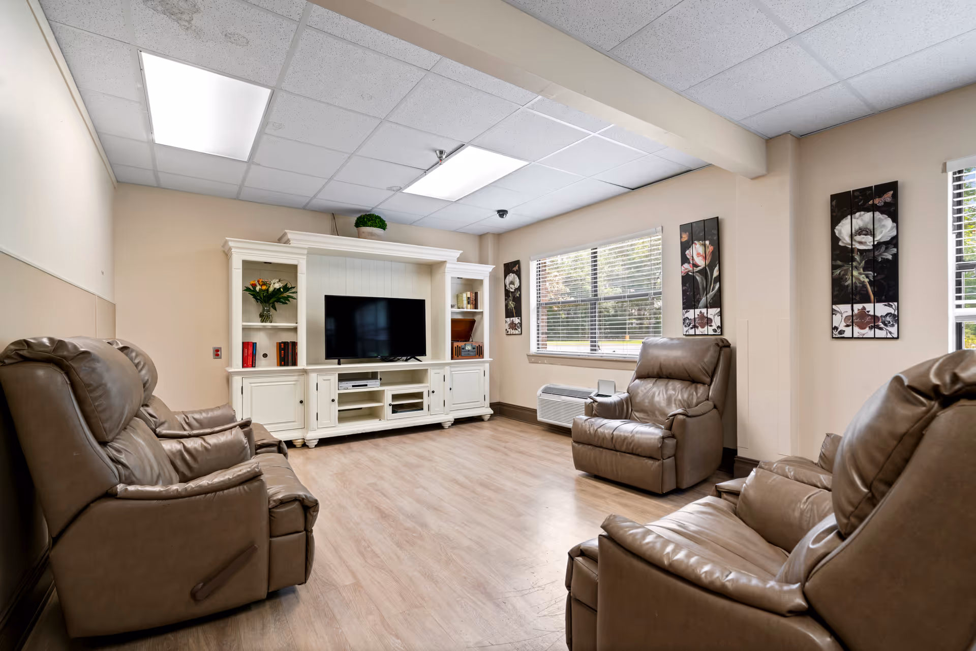 A cozy living room with four brown leather recliners arranged around a white entertainment center holding a flat-screen TV. The room has light-colored walls, a large window with blinds, floral artwork on the walls, and wood-look flooring.