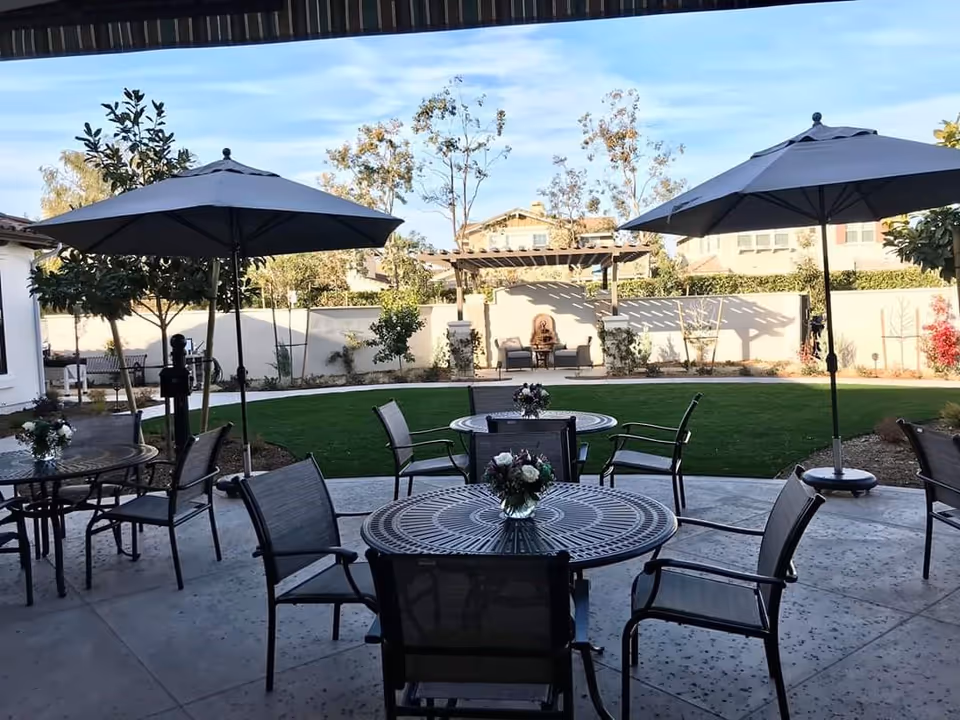 Outdoor patio area with round metal tables and chairs, each table decorated with a small flower arrangement. Two large umbrellas provide shade. Beyond the patio is a well-maintained lawn with trees and shrubs, and a pergola with seating against a white fence.