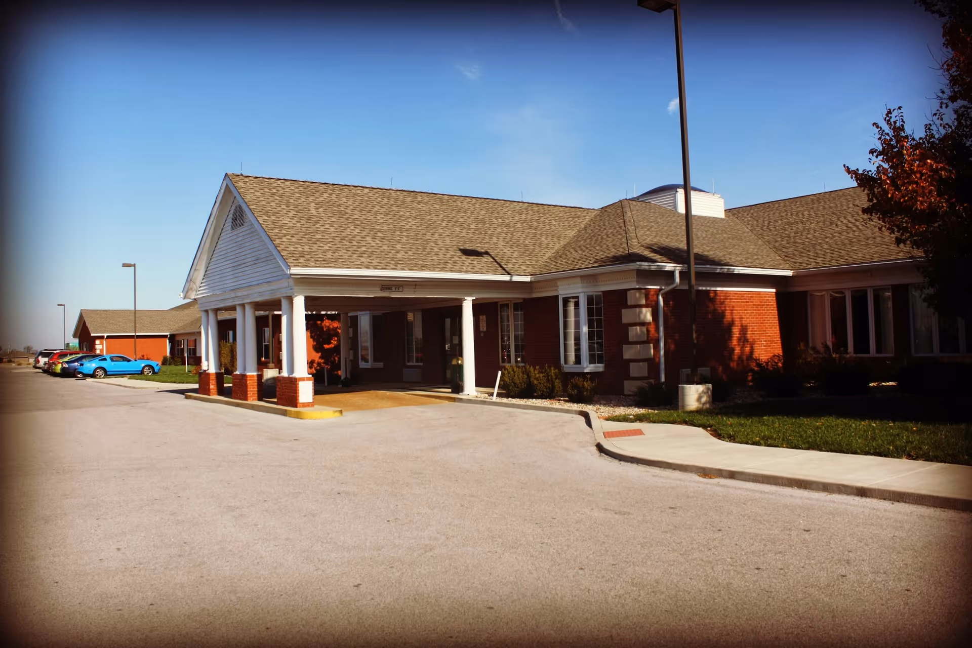 Exterior view of a single-story brick building with a covered entrance supported by white columns. Several cars are parked along the driveway, and there is a clear blue sky above.