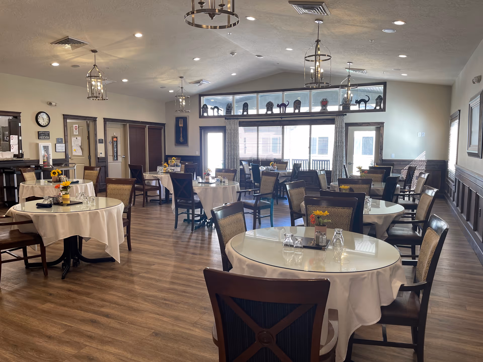 A dining room with several round tables covered with white tablecloths, each surrounded by wooden chairs with cushioned seats. The tables have small flower arrangements and glassware. The room has wooden flooring, large windows with blinds, and decorative items on a high shelf near the ceiling. Ceiling lights hang from above, and there is a clock and some wall decorations on the left side.