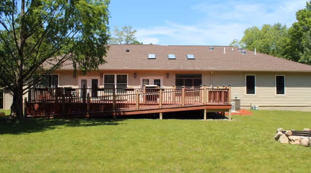 Single-story building with beige siding and a brown roof featuring skylights. A wooden deck with railings extends from the building, surrounded by a grassy lawn and trees under a blue sky.