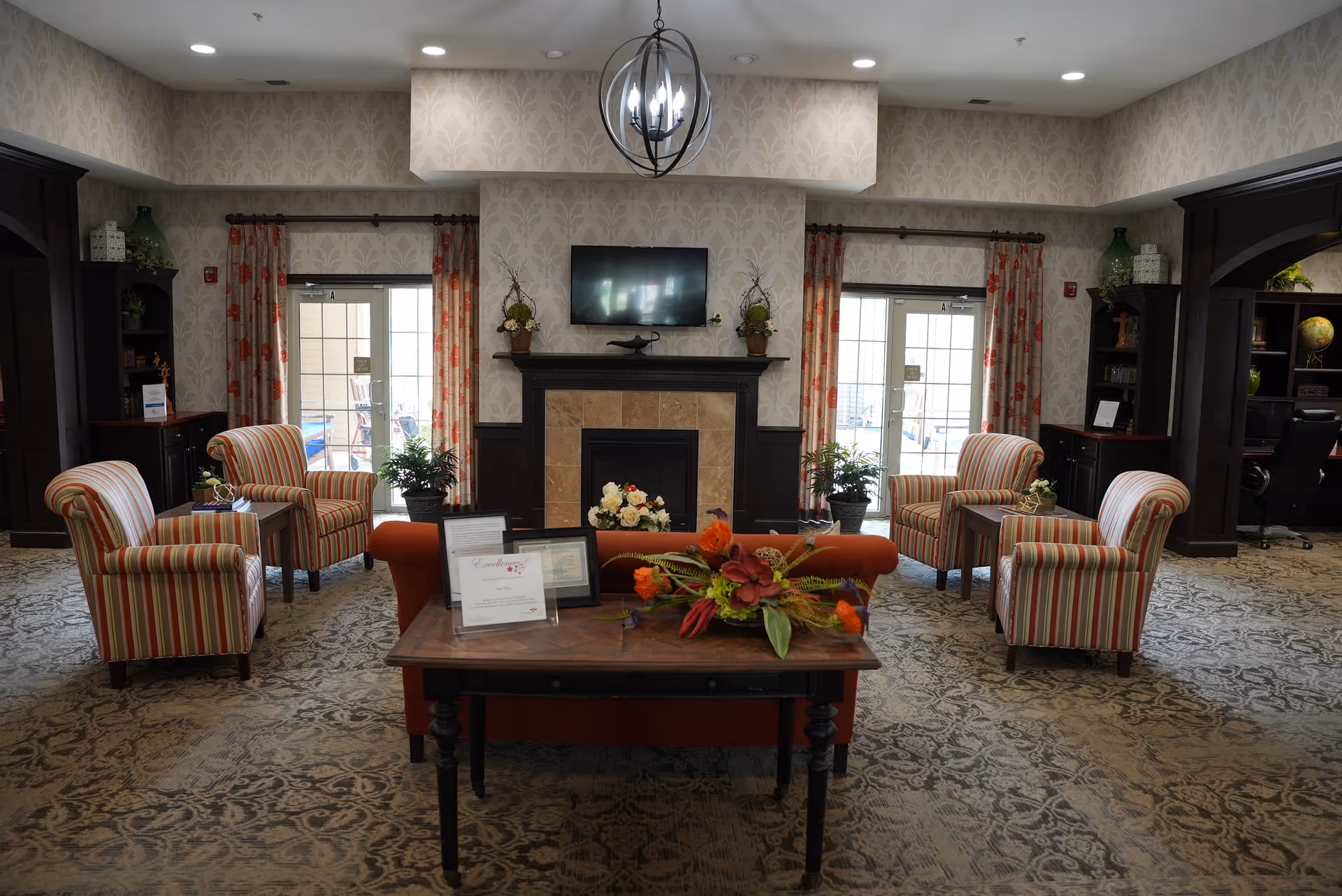 A cozy living room area in a senior living facility with a fireplace and a mounted TV above it. The room features four striped armchairs arranged around small side tables, two large windows with floral curtains, and a wooden table with flower arrangements and framed certificates in the foreground. The room has patterned carpet and wallpaper with a subtle floral design, and dark wood shelving units on either side.