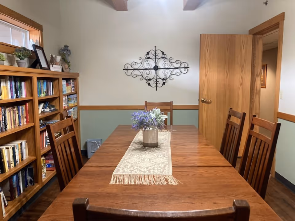 Wooden dining table with chairs and a floral centerpiece in a room with bookshelves and wall decor.