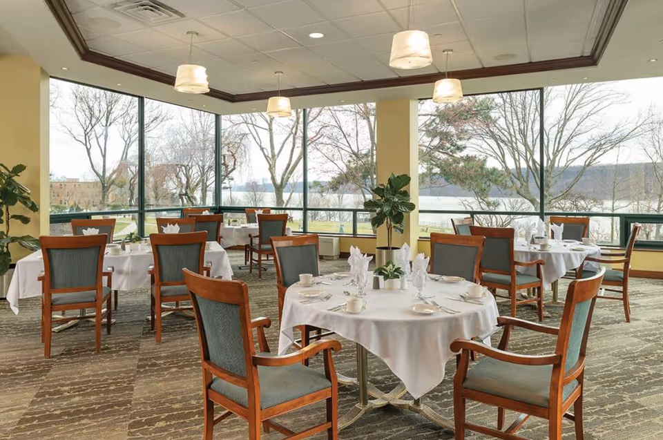 A bright dining room with several round tables covered with white tablecloths, each set with cups, plates, utensils, and napkins. The room has large floor-to-ceiling windows offering a view of trees and a body of water outside. Wooden chairs with green upholstery surround the tables, and there are ceiling lights and potted plants in the room.