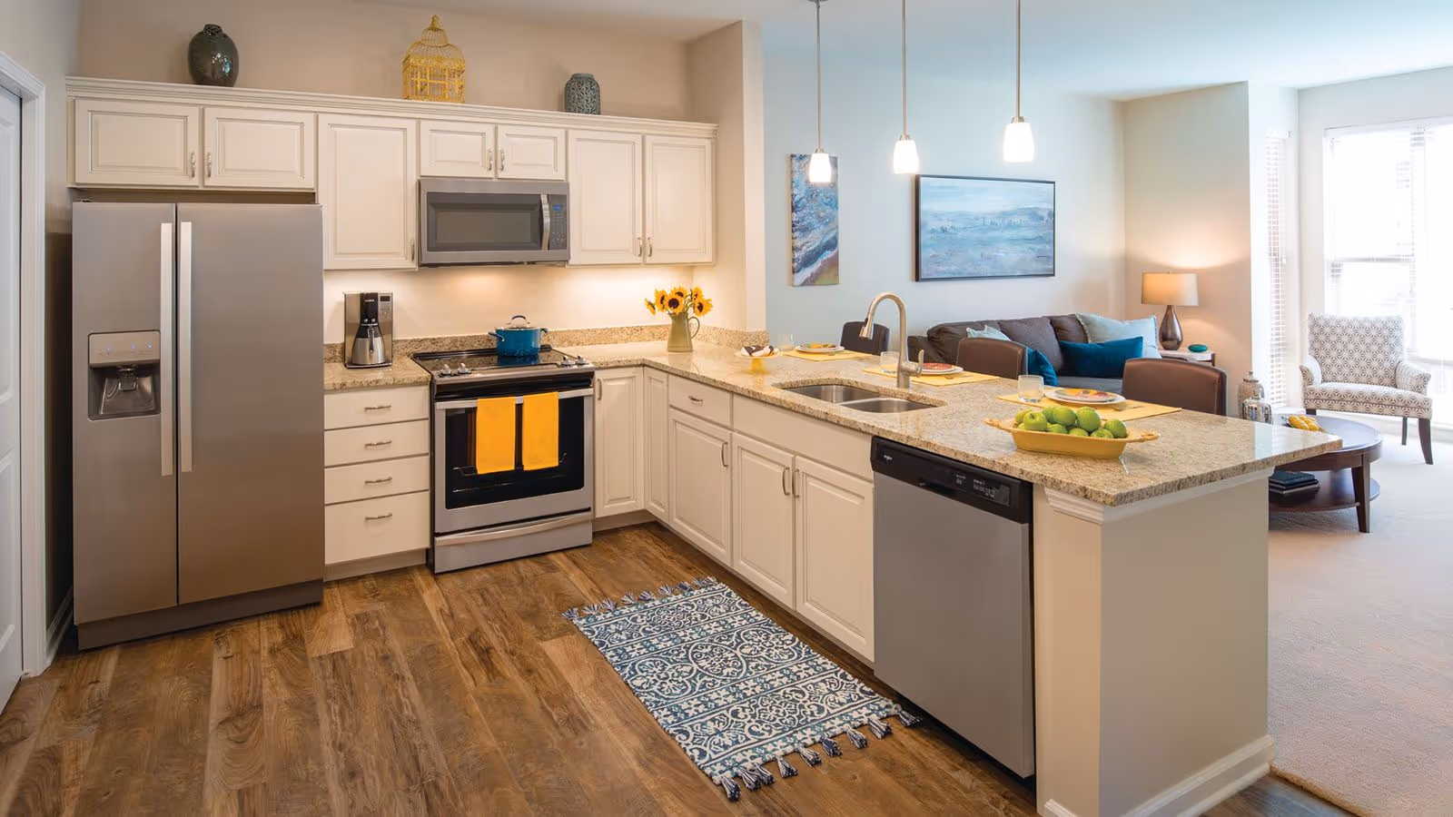 A modern kitchen with white cabinets, stainless steel refrigerator, oven, microwave, and dishwasher. The kitchen has a granite countertop island with a sink and seating area. The floor is wood, and the kitchen opens into a living room with a sofa, armchair, and large windows letting in natural light.
