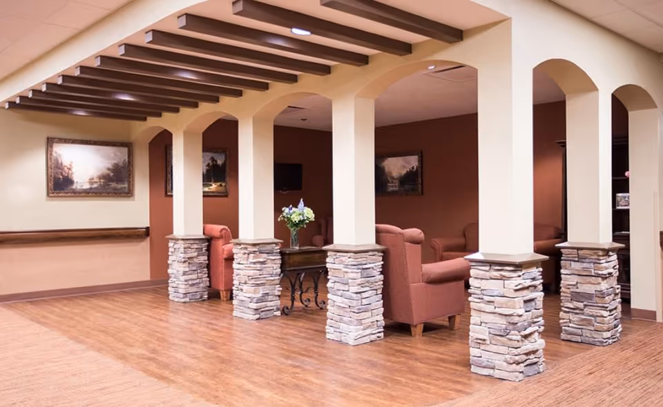Interior view of a senior living facility lounge area with wooden flooring, beige walls, and decorative stone pillars supporting arches. The room features several upholstered armchairs in a reddish color, a small table with a flower vase, and framed landscape paintings on the walls.