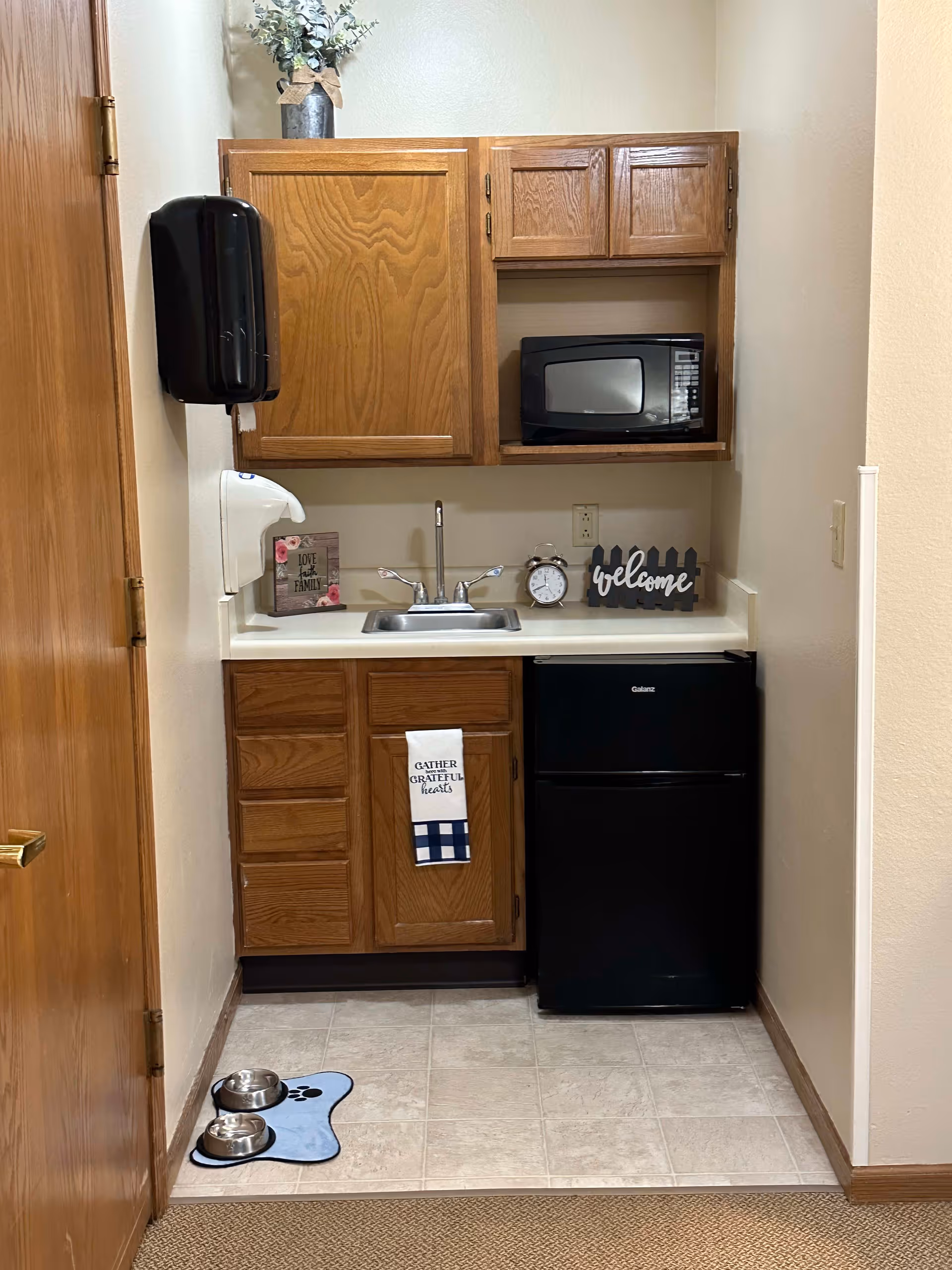 A small kitchenette area with wooden cabinets, a stainless steel sink, a black microwave on the upper shelf, and a black mini refrigerator below the counter. There is a decorative 'welcome' sign, a clock, and a small framed sign on the countertop. A towel with the text 'Gather Grateful Hearts' hangs from the cabinet door. On the floor, there is a blue pet feeding mat with two stainless steel bowls. A black wall-mounted soap dispenser and a white paper towel dispenser are on the left wall. The walls are painted light beige.