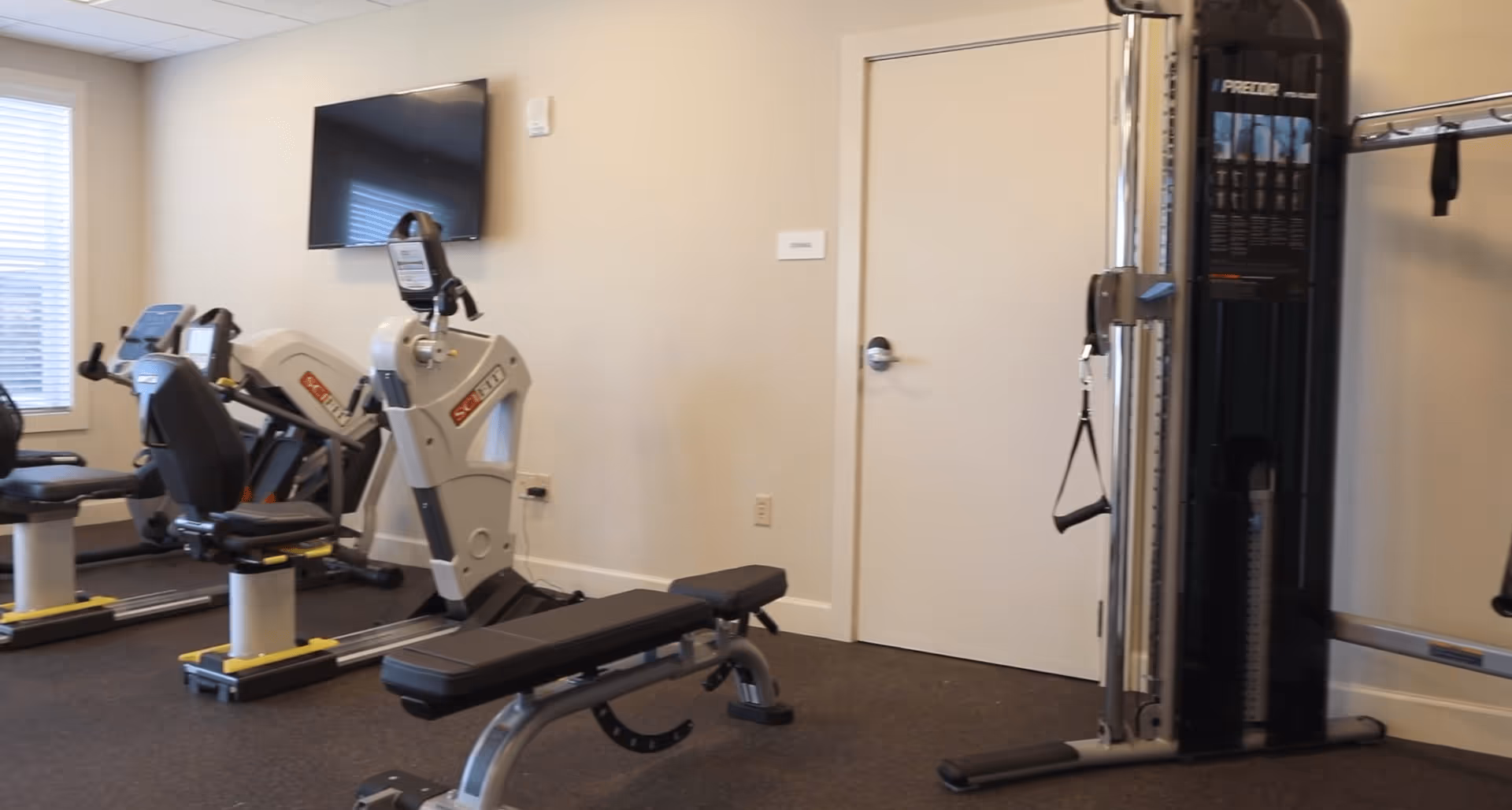 Interior view of a fitness room with exercise equipment including stationary bikes, a weight bench, and a multi-functional gym machine. A flat-screen TV is mounted on the wall, and there is a window with blinds letting in natural light.