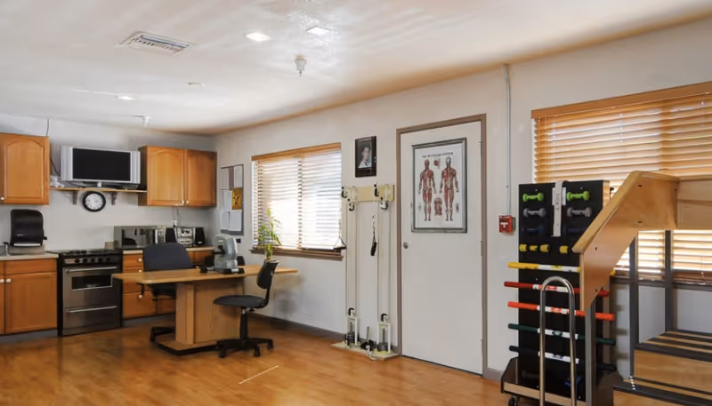 A room with a kitchen area featuring wooden cabinets, a stove, microwave, and a desk with an office chair. There are windows with blinds, a wall-mounted anatomical chart, and exercise equipment including weights and a wooden ramp.