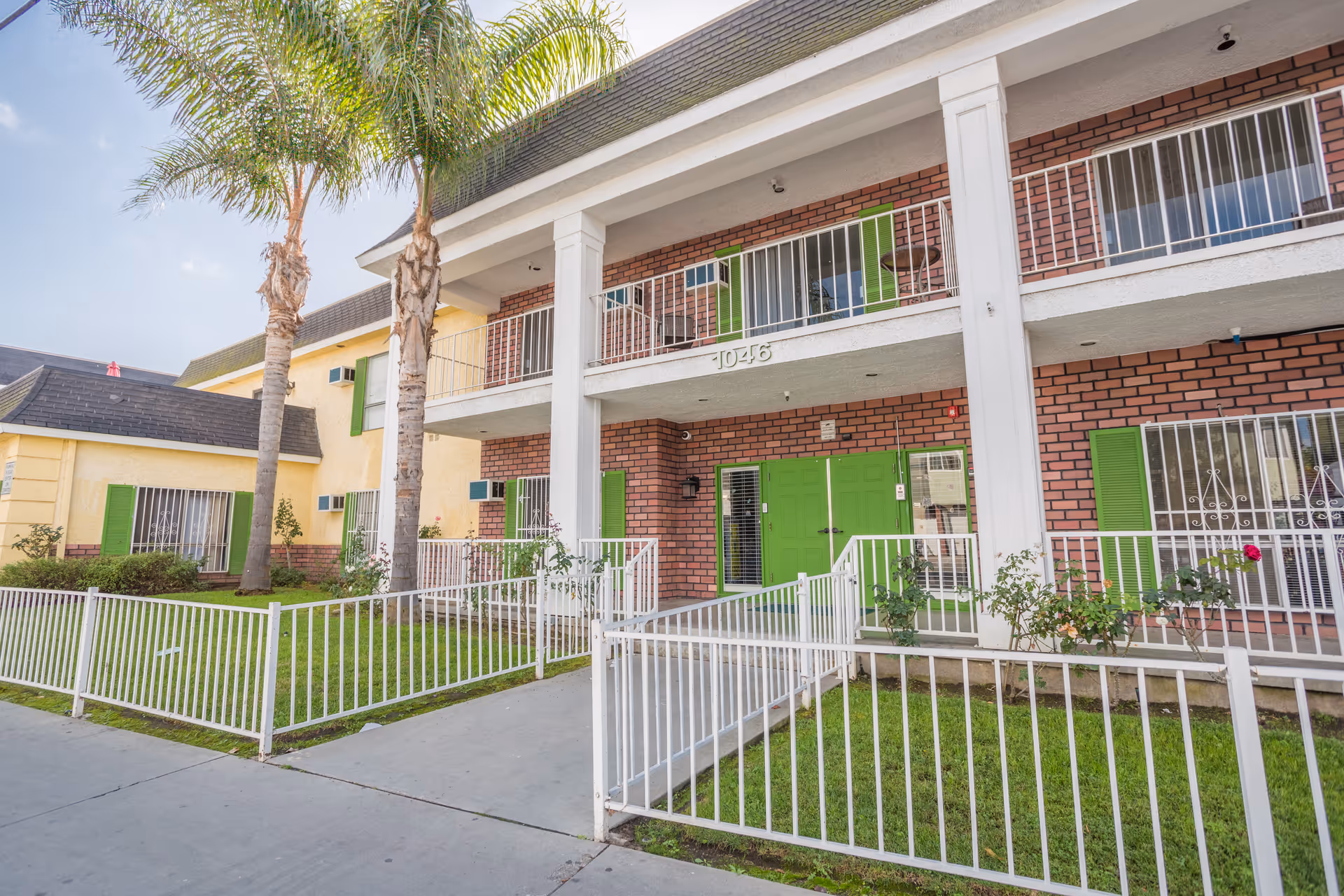 Exterior view of a two-story brick and yellow building with green doors and window shutters, white railings, and two palm trees in front. The building has the number 1046 displayed above the entrance.