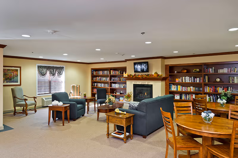 Cozy common room with sofas and chairs arranged around a fireplace, bookshelves and wooden dining tables.