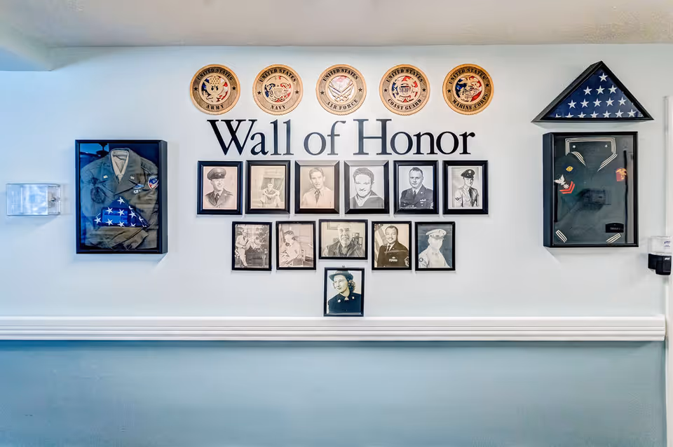 A Wall of Honor display featuring framed black and white photographs of military personnel, military insignias of the United States Army, Navy, Air Force, Coast Guard, and Marine Corps above the text 'Wall of Honor'. On either side of the text are framed military uniforms and folded American flags in triangular display cases.