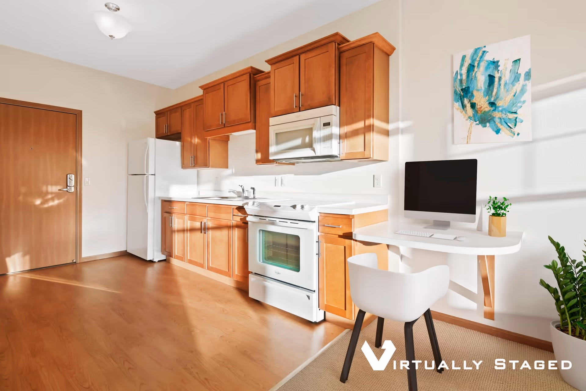 A bright kitchen area with wooden cabinets, a white refrigerator, stove, and microwave. To the right, there is a small white desk with a computer monitor, keyboard, mouse, and a small plant. A modern white chair is positioned at the desk. A colorful abstract painting hangs on the wall above the desk, and a green potted plant is on the floor nearby. The floor is wooden, and sunlight streams in, casting shadows on the walls and floor.