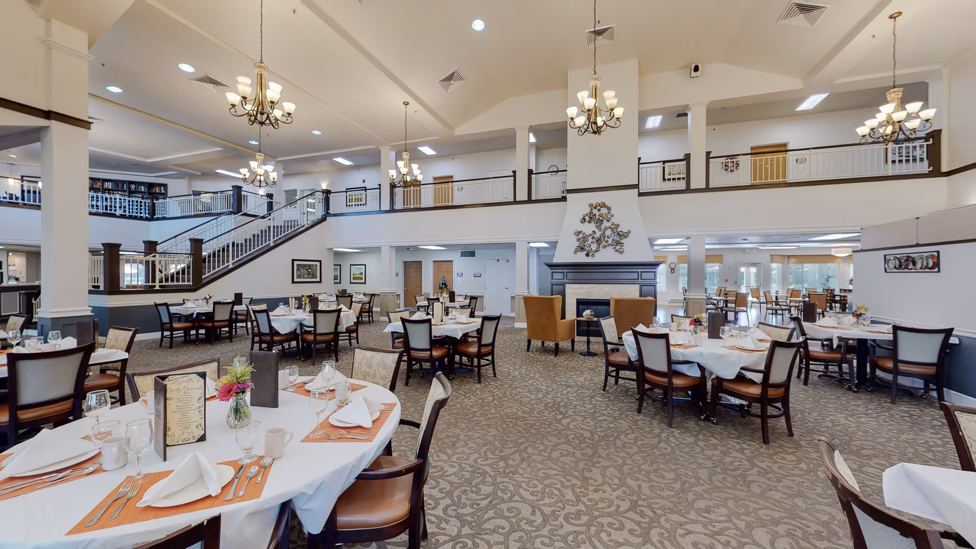 Spacious dining room with multiple round and rectangular tables set with white tablecloths, napkins, glassware, and silverware. The room features a high ceiling with chandeliers, a fireplace with two armchairs, and a second-floor balcony overlooking the area. The carpet has a patterned design, and the walls are decorated with framed artwork.