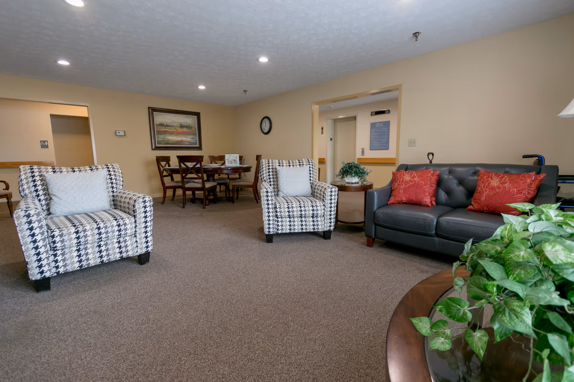 A cozy living room area in a senior living facility featuring two patterned armchairs with white pillows, a dark gray sofa with red pillows, a round wooden table with a green plant, and a dining table with chairs in the background. The walls are beige, and there is a framed painting and a clock on the wall.