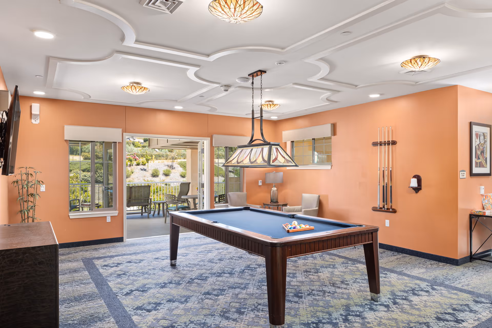 A recreational room with a pool table in the center, featuring a blue felt surface and a rack of billiard balls. The room has orange walls, a patterned carpet, and a decorative ceiling with multiple light fixtures. There are two beige chairs and a small table with a lamp in the corner. Large windows and a sliding door open to an outdoor patio area with chairs and greenery visible outside.