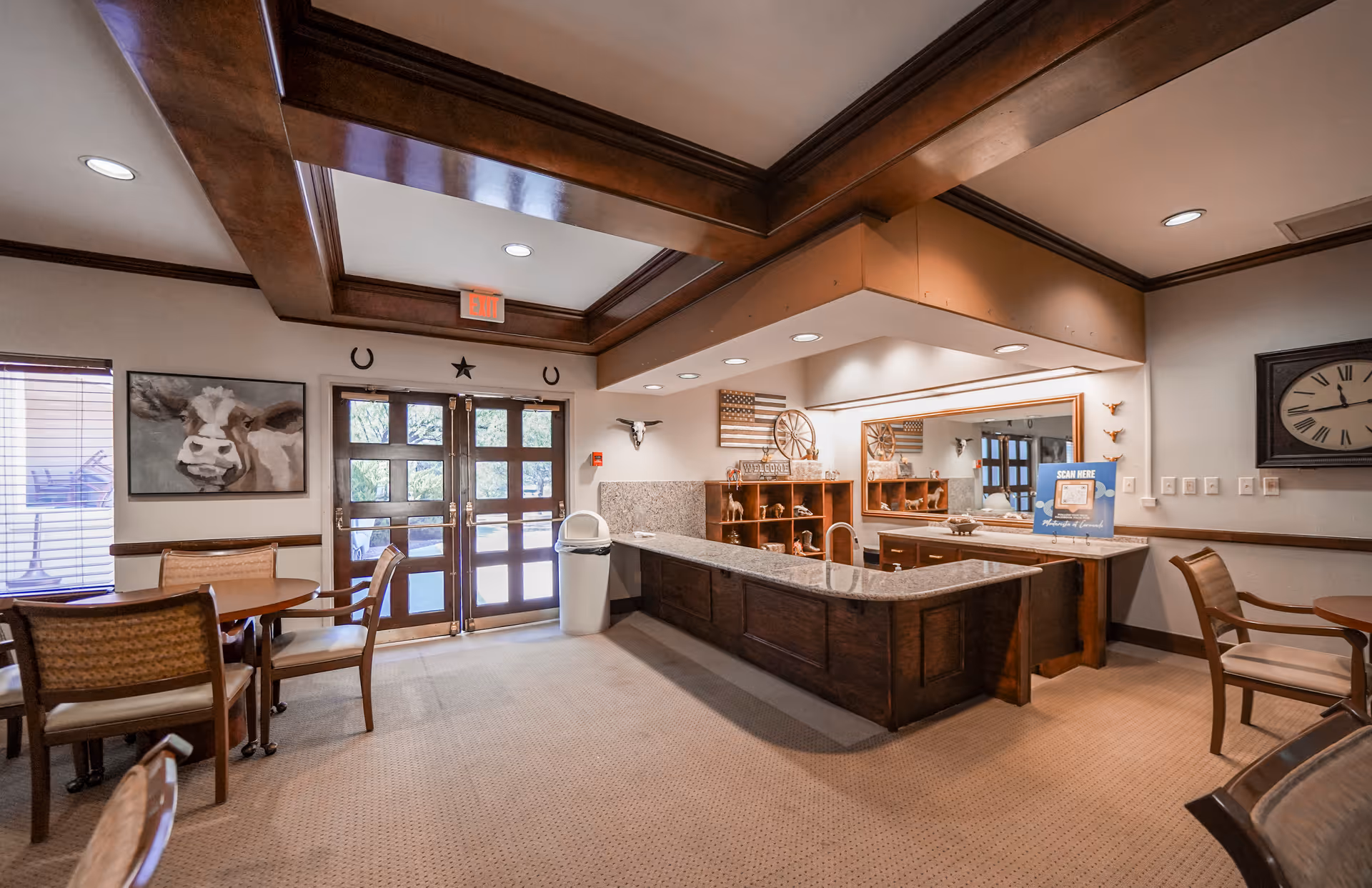 Interior view of a senior living facility common area with a wooden reception desk, round tables with chairs, a large wall clock, and decorative artwork including a cow painting and a wooden American flag. The room has a beige carpet, wooden ceiling beams, and double glass doors leading outside.