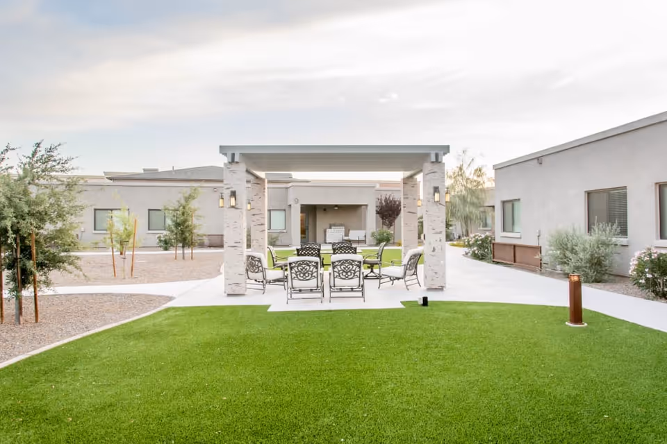 Outdoor seating area with a covered pergola featuring a table and several chairs on a concrete patio surrounded by green grass and landscaped plants, with single-story buildings in the background under a cloudy sky.