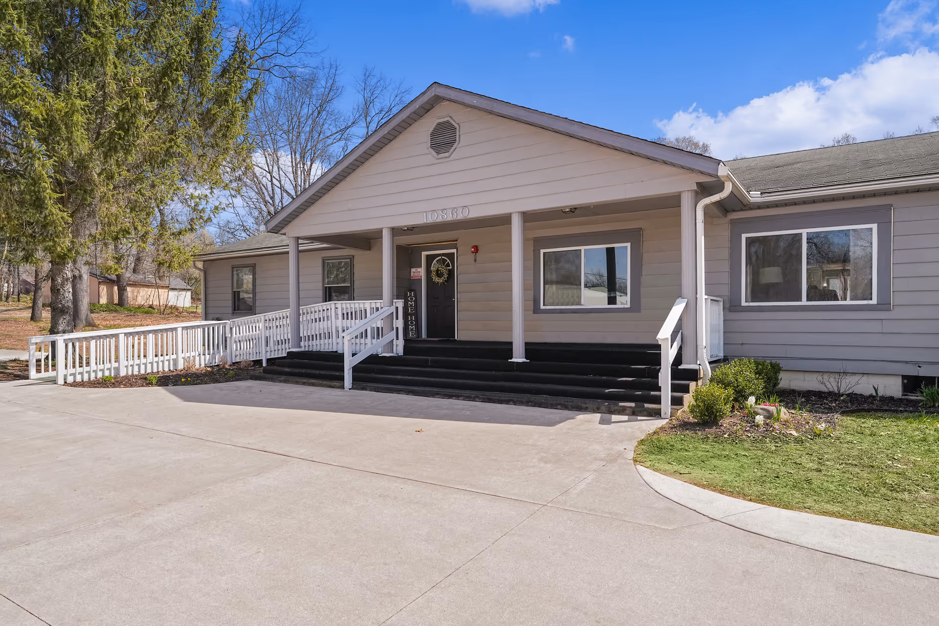 Exterior front view of a single-story assisted living facility building with a covered entrance, steps, and a wheelchair ramp. The building is light gray with white trim, surrounded by trees and a clear blue sky.