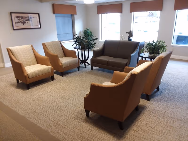 A cozy seating area in a senior living facility with four beige armchairs arranged around a dark brown loveseat. There are two small round tables with green potted plants on either side of the loveseat. The room has large windows with brown valances, beige carpet, and a framed picture on the wall.