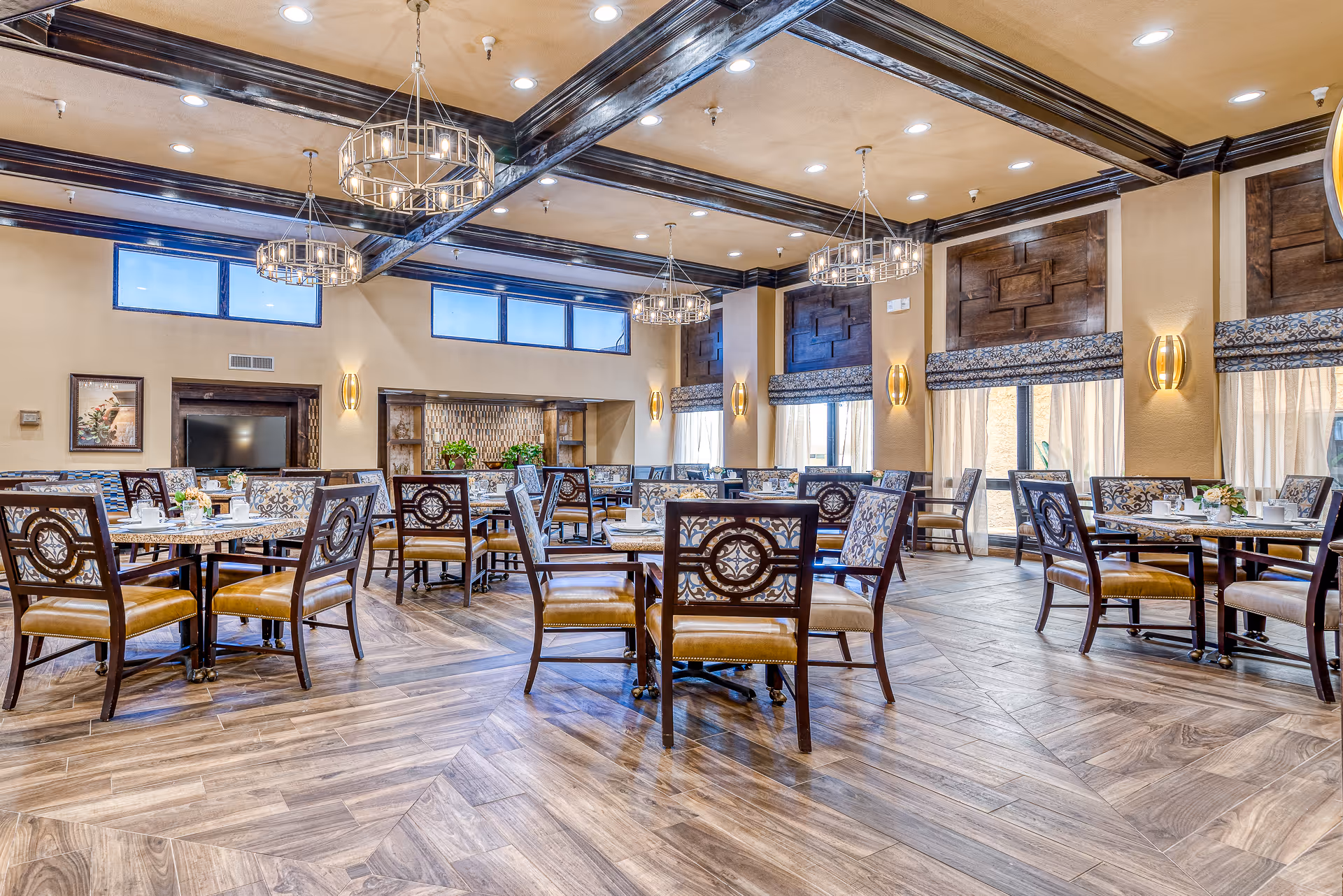 Spacious, elegant dining room with multiple set tables and chairs, decorative chandeliers, and exposed wooden ceiling beams.