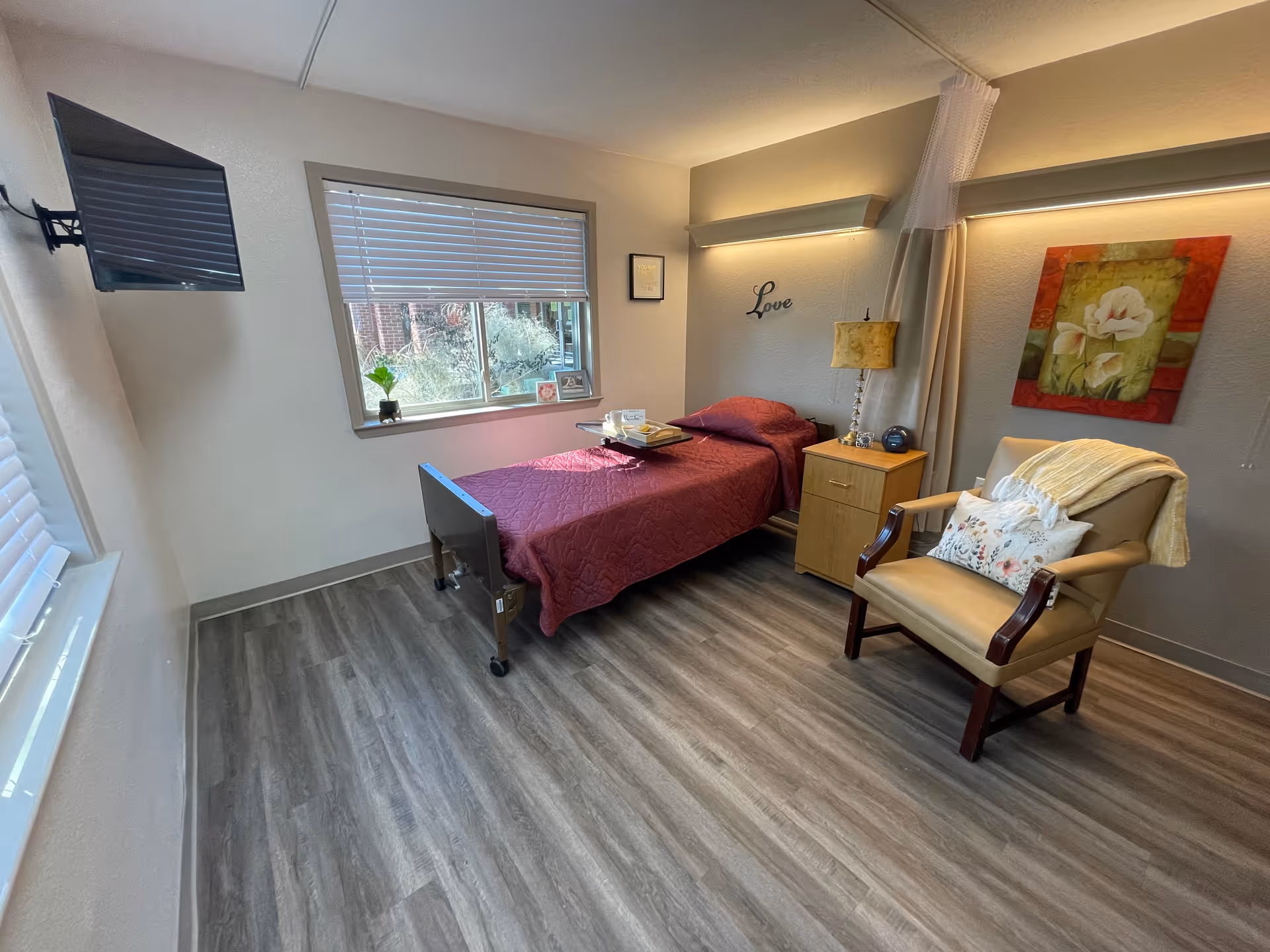 A bright, tidy senior living bedroom with a single bed, bedside table, armchair, wall art and a wall-mounted TV.