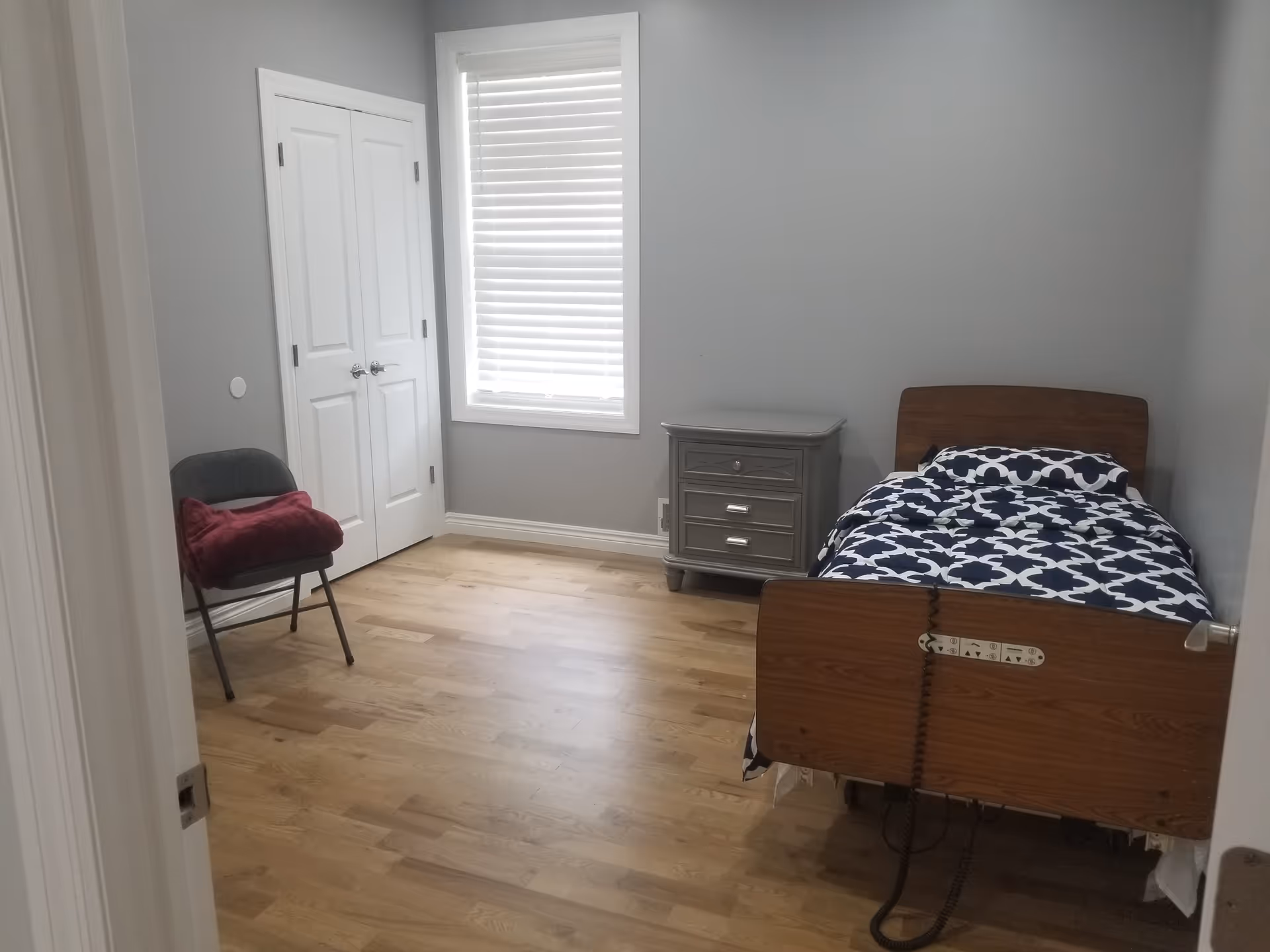 A simple bedroom with light gray walls and wooden flooring. The room contains a single hospital-style bed with a blue and white patterned blanket, a gray nightstand with drawers, a window with white blinds, a closed white double-door closet, and a gray folding chair with a red blanket draped over it.