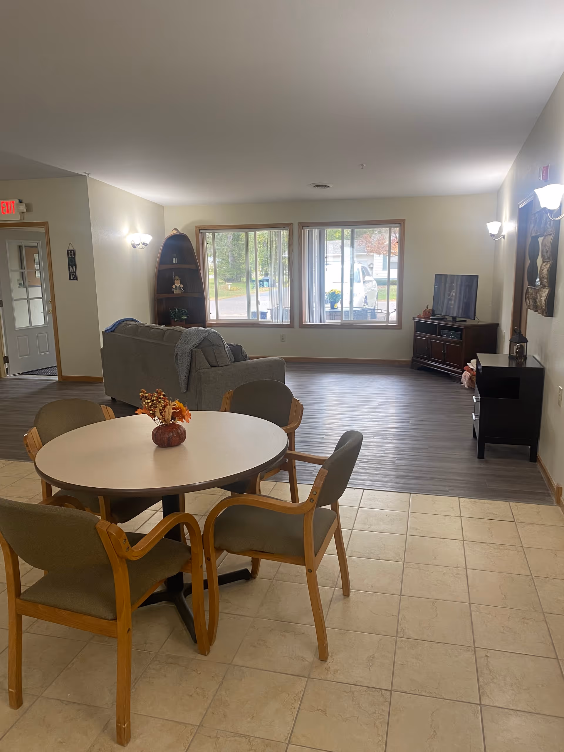 A senior living facility common area with a round table and four chairs in the foreground, a gray couch facing a TV on a stand in the background, large windows letting in natural light, and light-colored tiled and wood flooring.