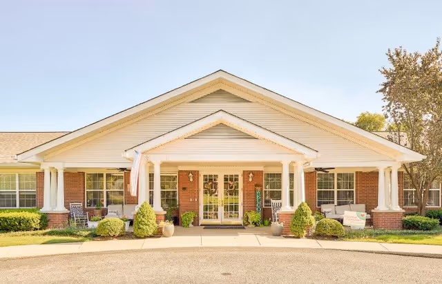 Front exterior view of a single-story brick building with white columns and a peaked roof. There are shrubs and small trees in front, a concrete walkway leading to double glass doors, and an American flag hanging near the entrance.