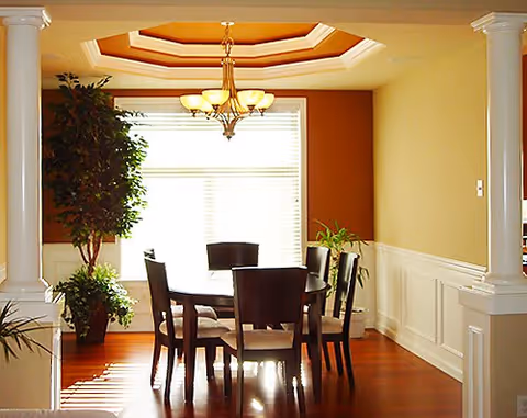 A dining room with a round wooden table surrounded by six chairs. The room features hardwood floors, a large window with blinds, a chandelier hanging from a recessed ceiling, and decorative white columns framing the space. There are also two potted plants near the window.