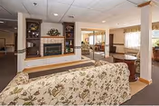 Interior view of a senior living facility common area featuring a floral-patterned sofa in the foreground, a fireplace with built-in shelving on either side, and a large mirror reflecting additional seating and windows with curtains. The room has a carpeted floor and a drop ceiling with recessed lighting.